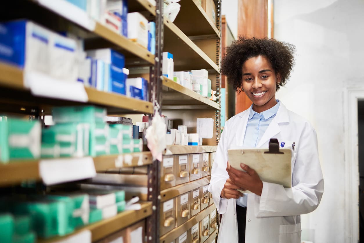 Confident African female pharmacist standing by a medicine shelf holding a clipboard.